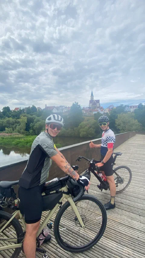 Both riders posing with their bikes on a wooden bridge, the town's skyline behind