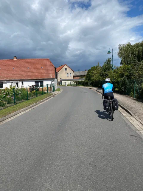 Rider in a light-blue jersey on a village road, red roofs and dark clouds behind