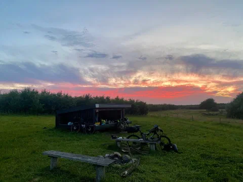 Black wooden shelter with the bikes and a bench on a meadow, pink-orange dusk sky