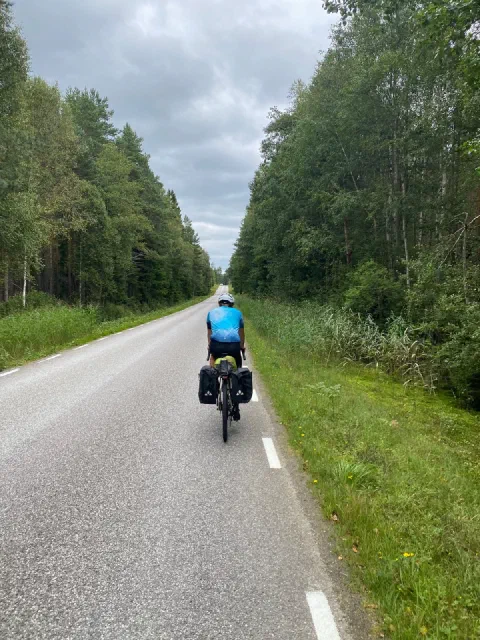 Rider in a light-blue jersey on a straight Swedish road through birch and spruce forest
