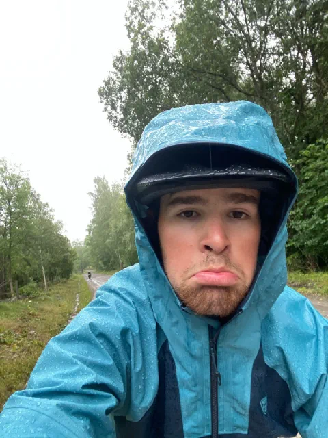 Grumpy selfie in a turquoise rain jacket and helmet, a wet road behind
