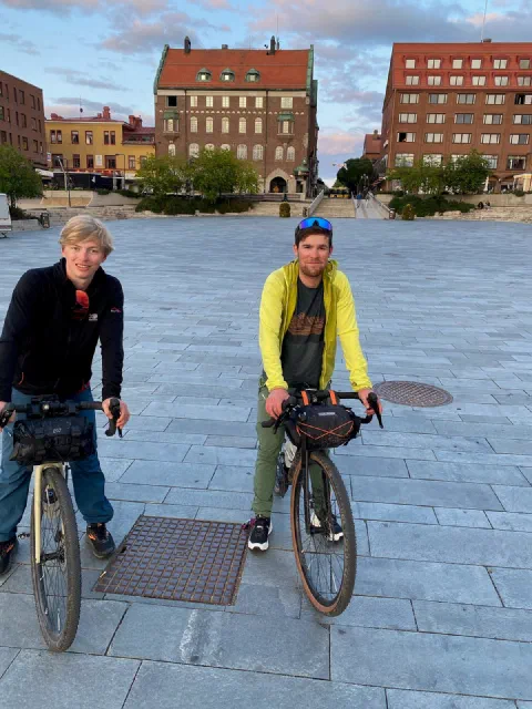 Both riders with their bikes on a paved square in Östersund, red-brick buildings behind