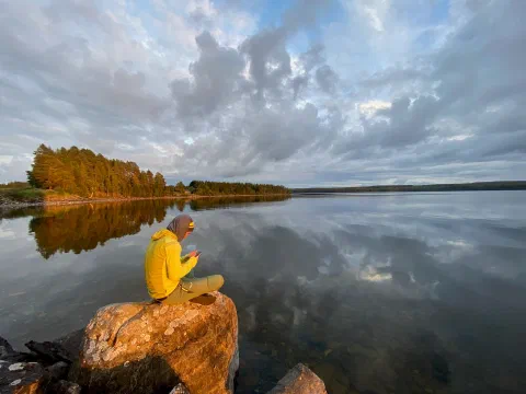 Friend in a yellow jacket on a sunlit boulder by a still lake, golden trees across