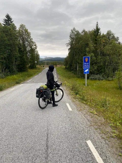 Rider with a loaded bike beside the Norwegian border sign on an empty road, birch forest behind