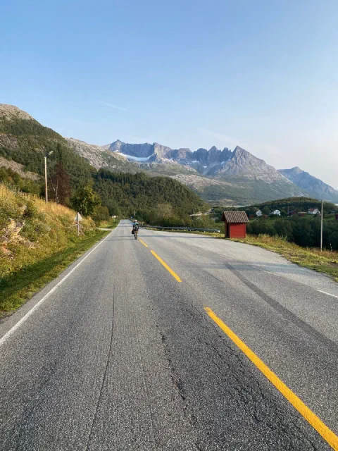 Rider far down a wide road towards sharp snow-capped peaks, a red hut in the evening light