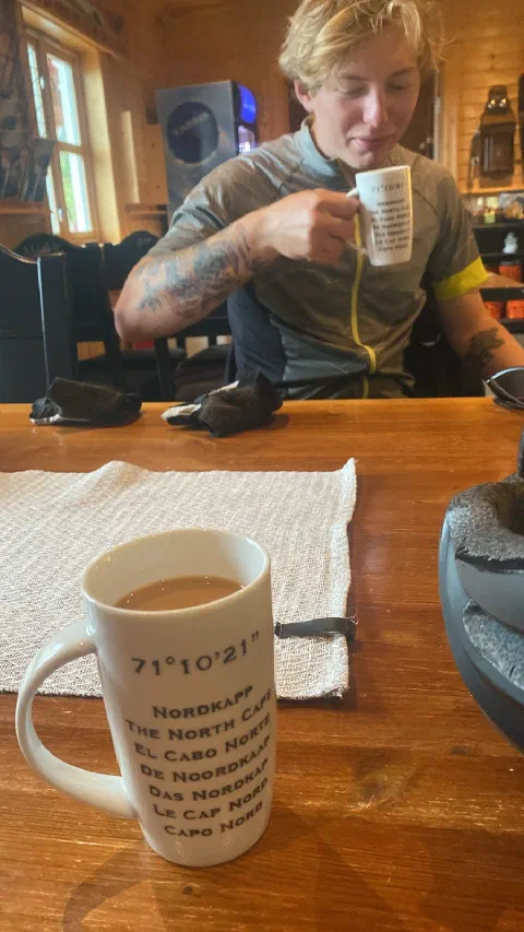 Rider drinking from a "Nordkapp 71° 10' 21" mug in a wooden café