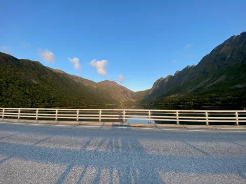 Bridge railing with long shadows, green mountains under a blue sky
