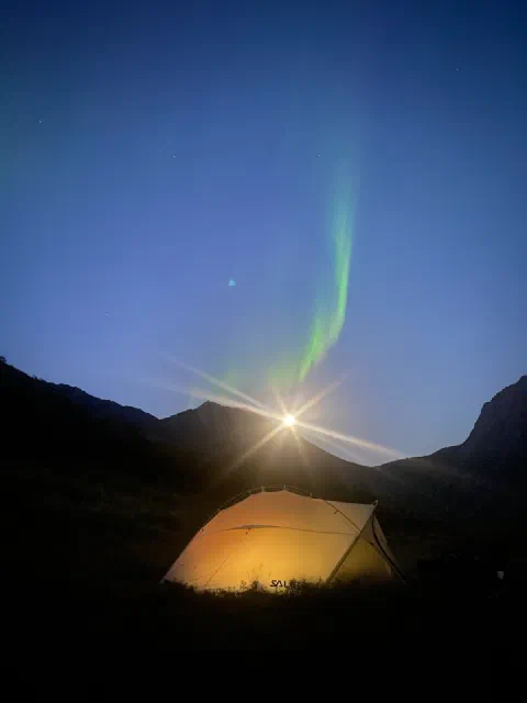 Northern lights over a glowing yellow tent, the moon beside the ridge