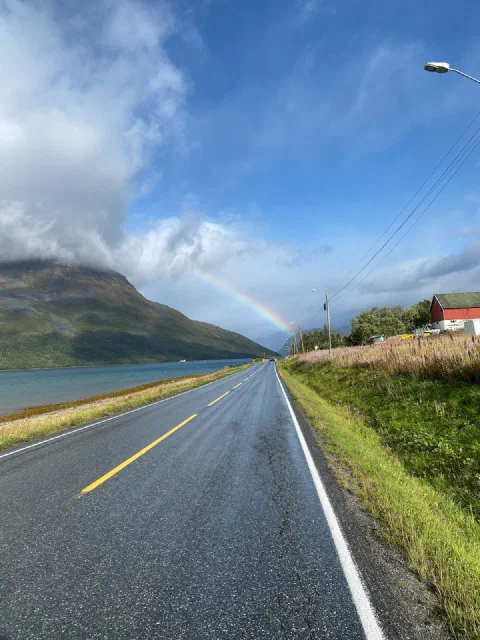 A rainbow over a fjord and a wet road, a red house by the verge