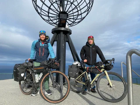 Both riders with loaded bikes at the Nordkapp globe under a dramatic sky