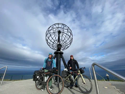 Both riders with their loaded bikes under the Nordkapp globe, streaky clouds