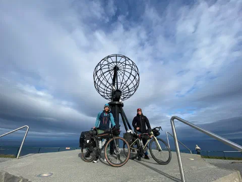 Both riders at the Nordkapp globe, the sea horizon behind and a visitor at the railing