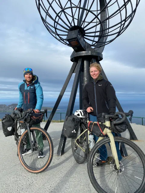 Both riders smiling at the Nordkapp globe with their loaded bikes, blue sky