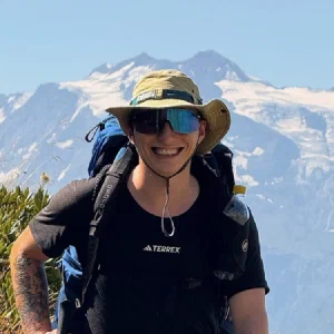Max hiking with a backpack and sun hat, smiling, in front of glaciers